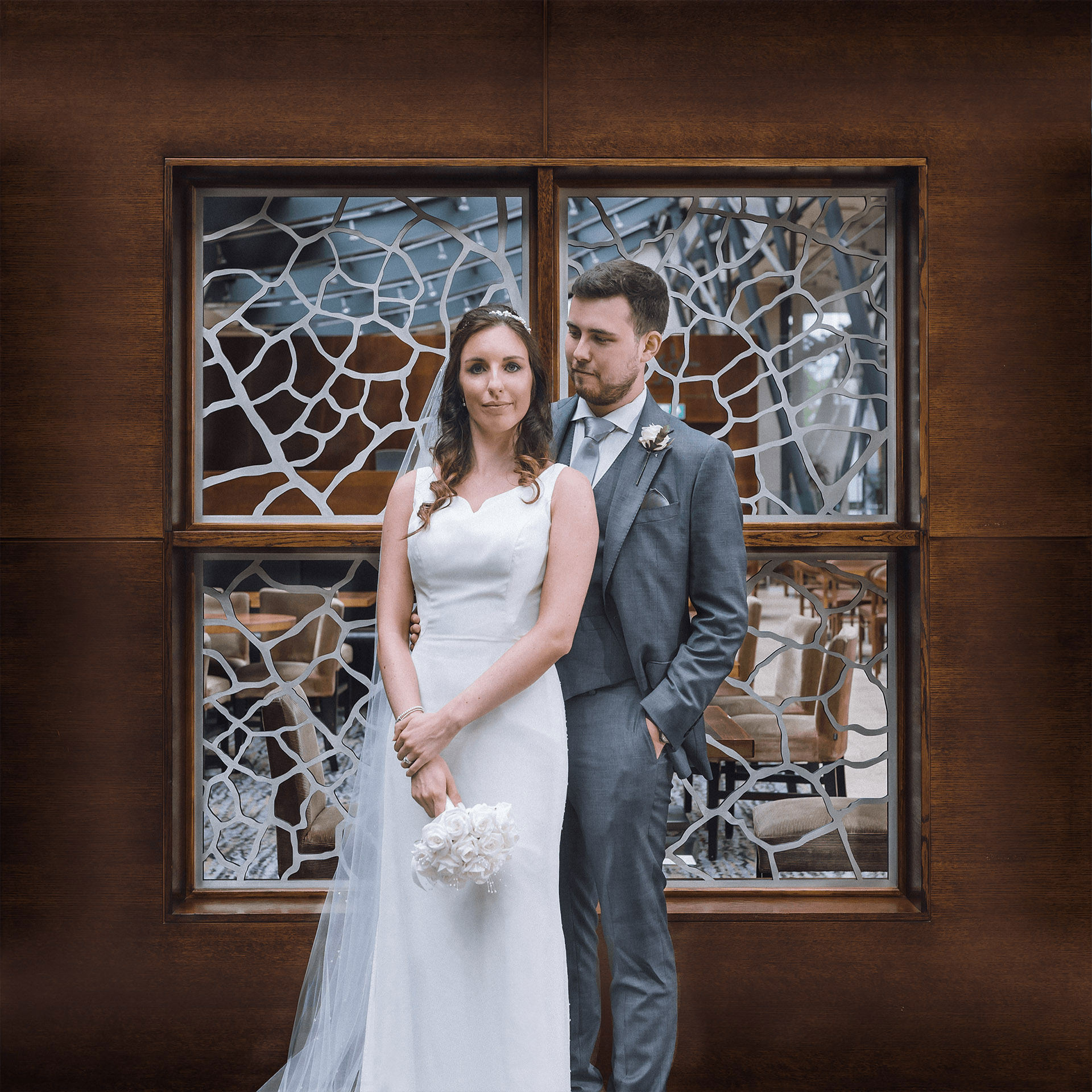 The bride and groom stand together in front of a square wooden panel