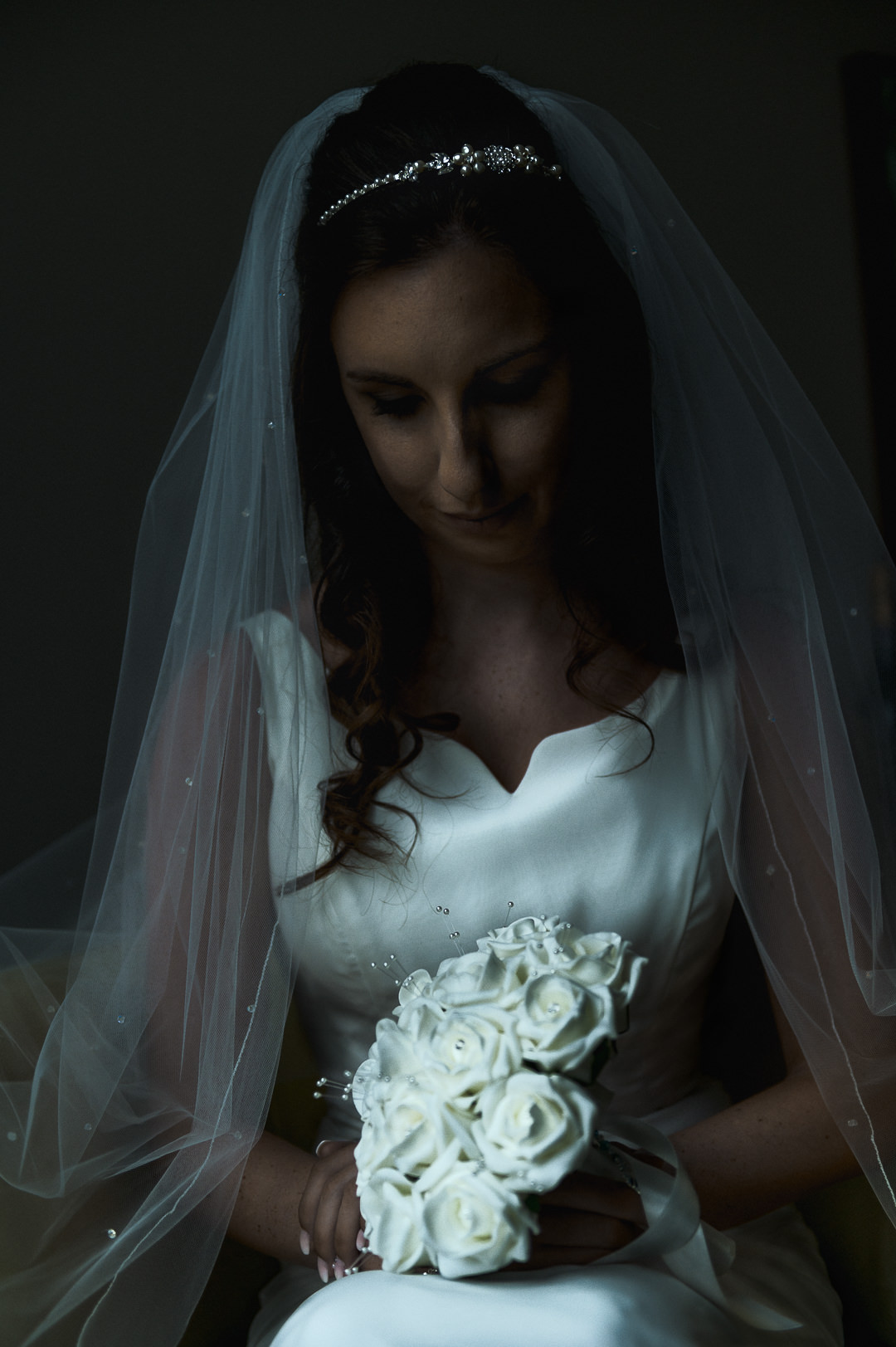 The bride is sitting and holding her flowers which are bright in the window light