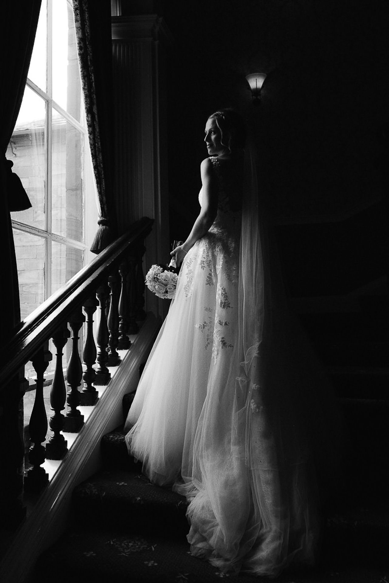 black and white photo of bride on the stairs looking out of the window at walton hall