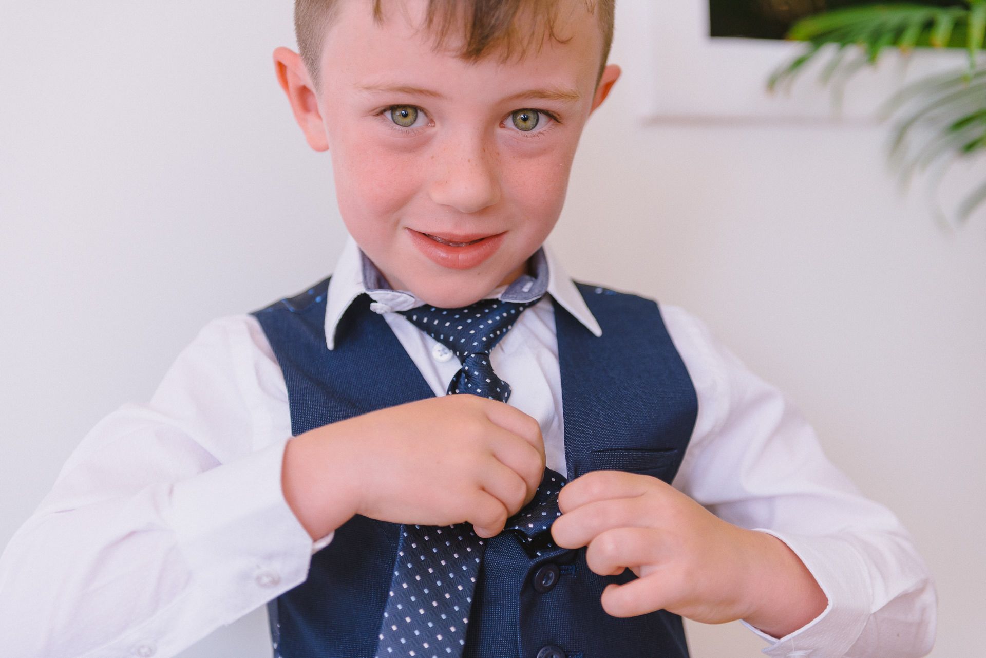 a young pageboy smiles as he tucks his tie into his waistcoat