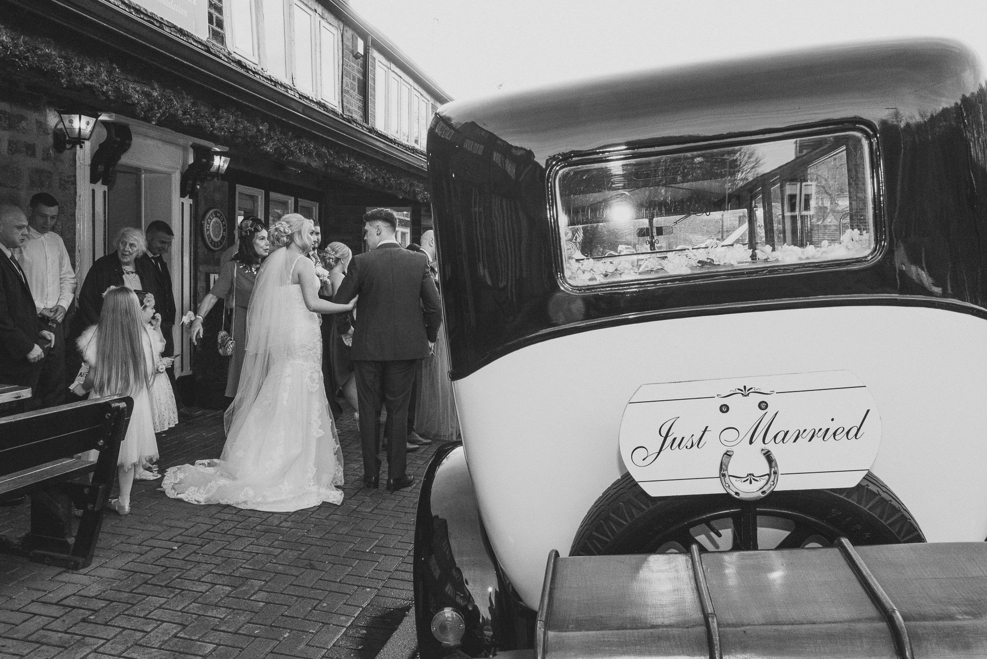 view of back of wedding car with bride and groom stood to the side